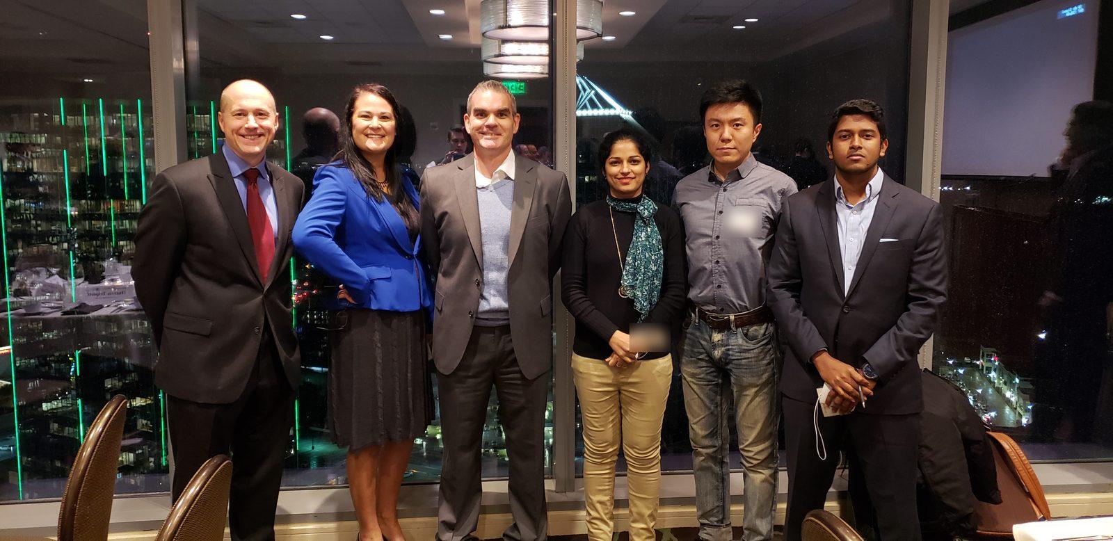 Six members of the hackathon team standing side by side at the finalist dinner, with city lights through the windows behind them.